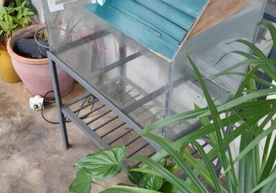 Miniature blue house model with a sloped roof and small balcony, displayed on a glass stand.