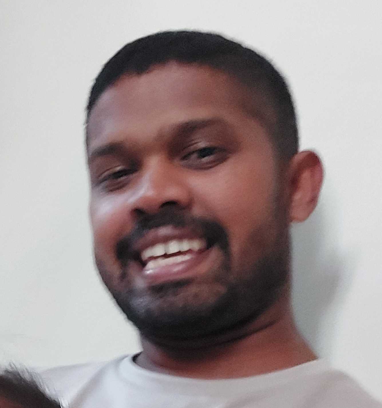 Happy man with a broad smile, wearing a light grey casual T-shirt, posing indoors against a plain white wall.