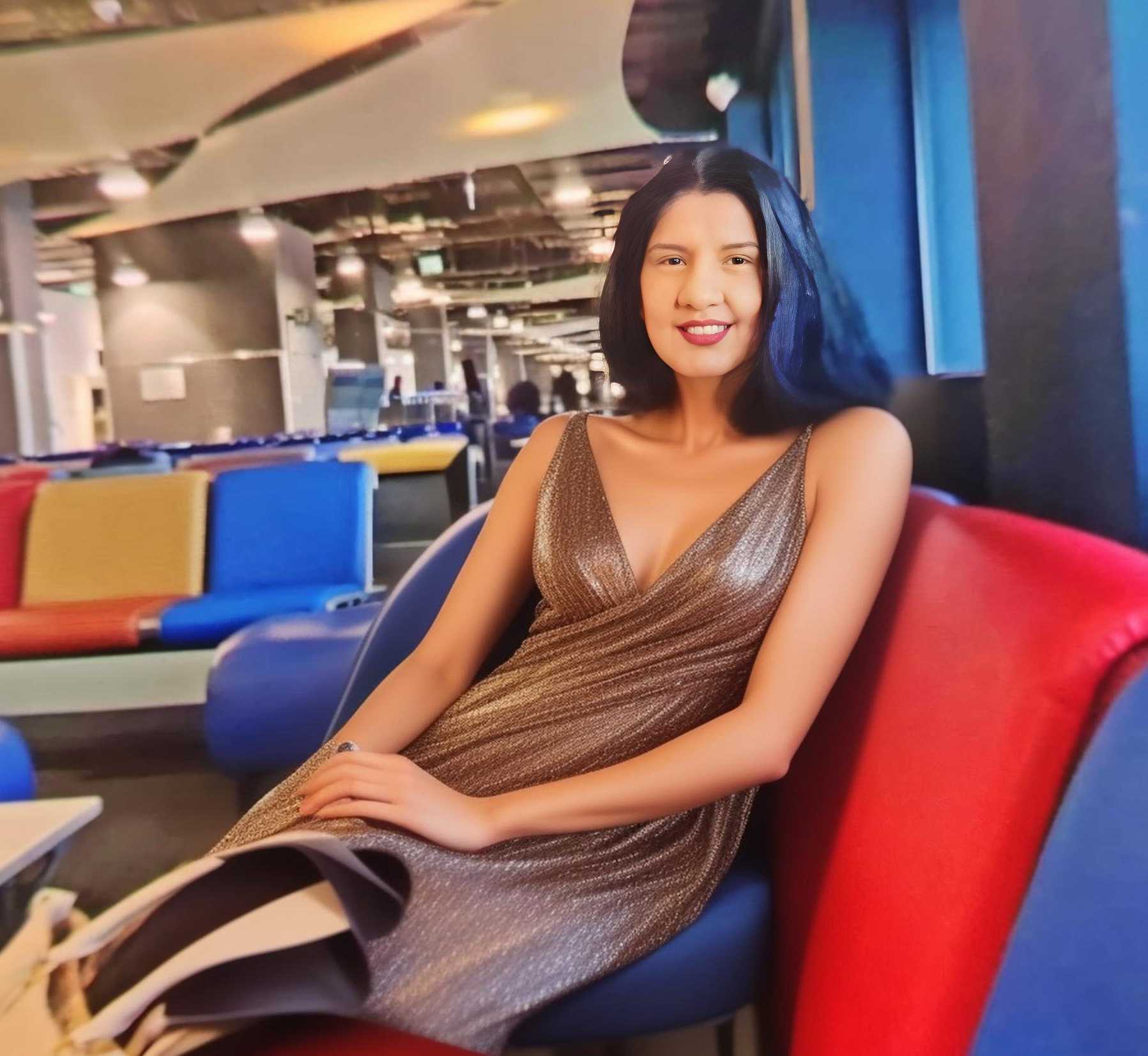 Beautiful woman with black hair in a shiny gold dress, sitting comfortably in a vibrant lounge area.