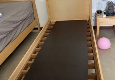 Wooden bed frame with black mattress, light wood finish, in a modern bedroom setting.