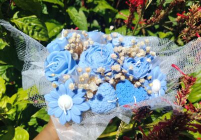 Blue floral bouquet with roses and daisies, wrapped in tulle, placed outdoors among green foliage.
