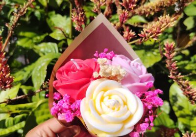 Beautiful flower bouquet with pink, white, and red roses, wrapped in pink paper with a ribbon, held outdoors among lush greenery and flowering plants.